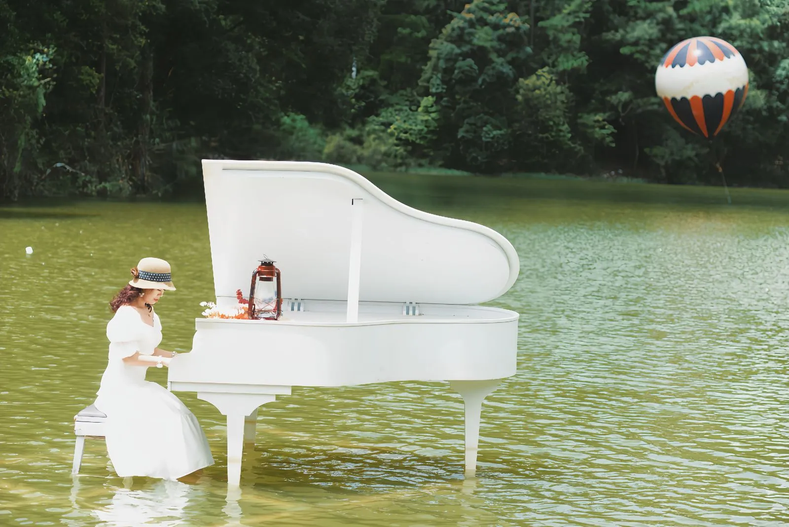 A woman plays a white grand piano in a serene lake setting with a hot air balloon in the background.